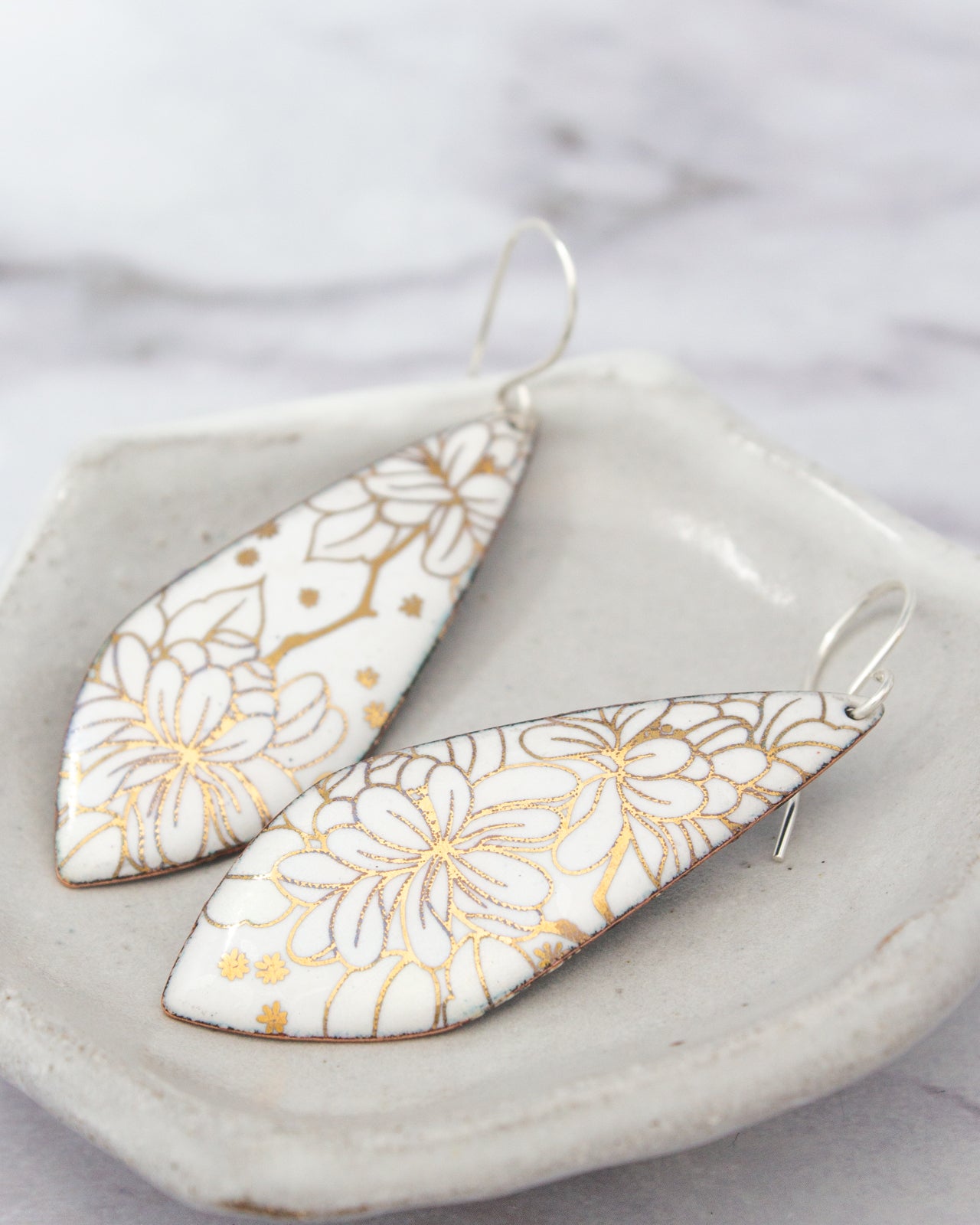 two white earrings with gold floral designs, placed on a white plate or tray.