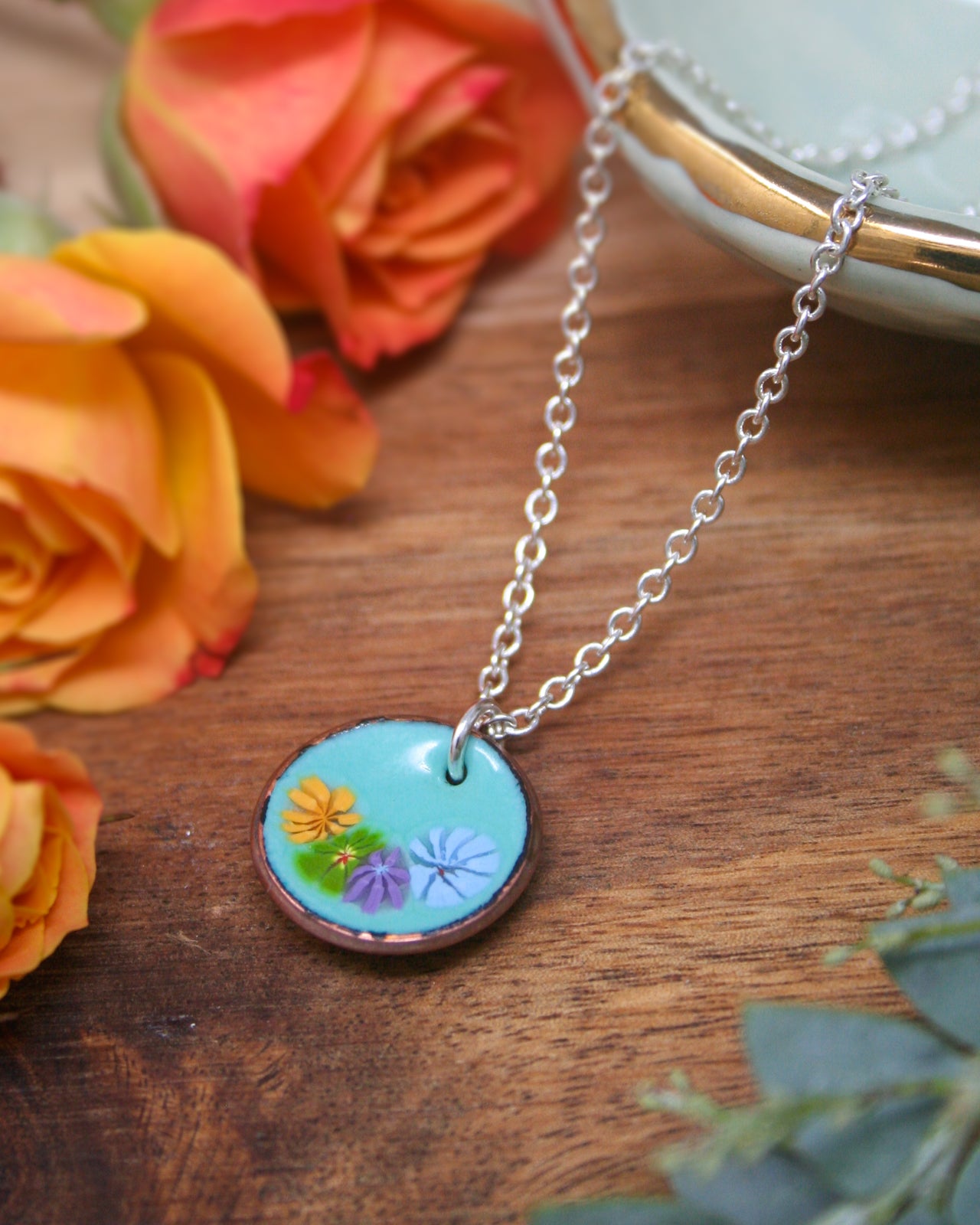 A necklace with a pendant featuring a floral design is displayed on a wooden surface, surrounded by vibrant orange and yellow roses.