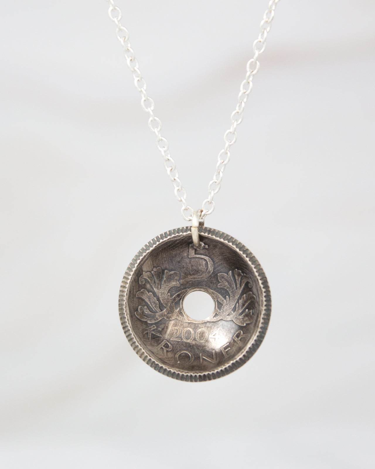 A silver necklace pendant with an embossed design, featuring a hole in the center surrounded by decorative elements.