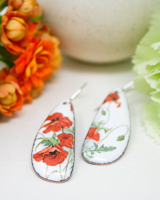 a pair of earrings with a floral design, including red flowers and green leaves, placed on a white surface.