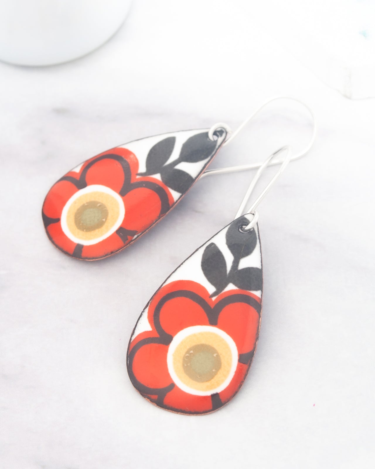 A pair of earrings with a floral design, featuring red and black elements, are displayed on a white surface.