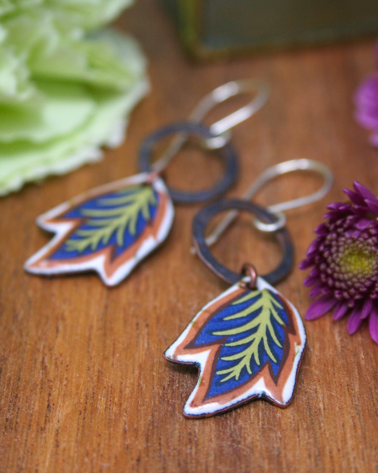 a pair of colorful earrings with leaf-shaped designs, placed on a wooden surface alongside a purple flower.