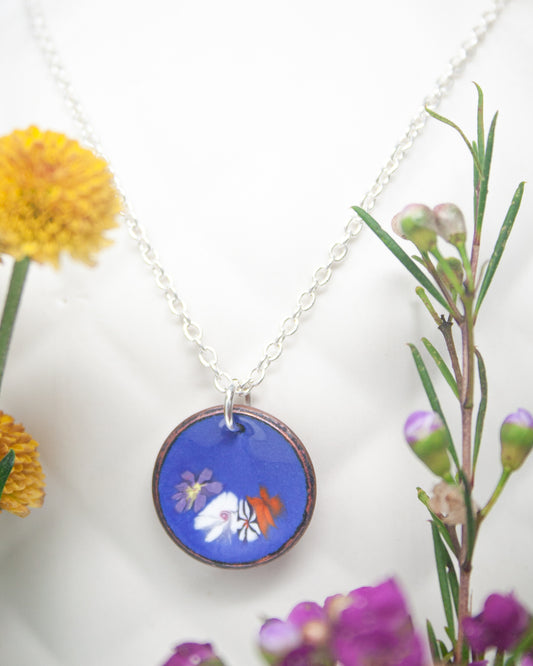 A necklace with a blue pendant featuring a floral design is displayed against a white background, with yellow flowers and purple flowers visible in the background.
