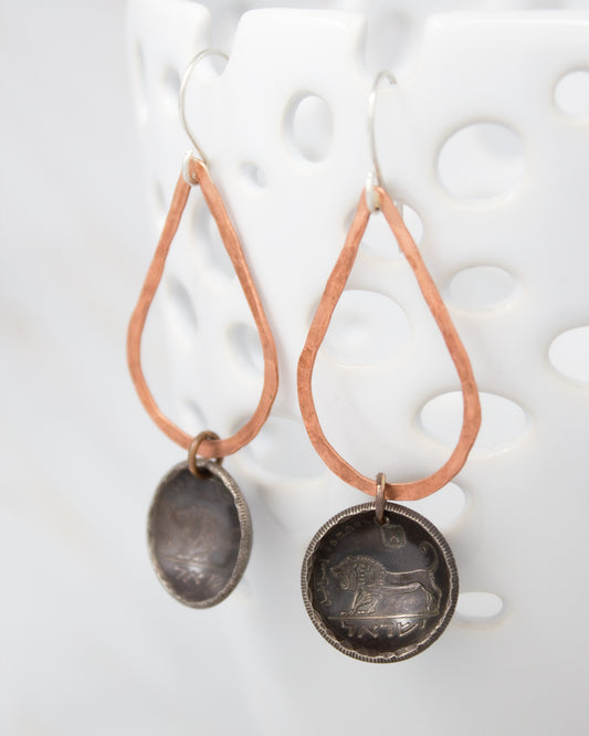 A pair of earrings with a metal lion design on the circular pendant, hanging from a copper-colored metal frame.