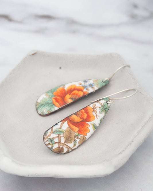 A pair of earrings with a floral design, featuring orange and green flowers, are displayed on a white plate against a blurred background.