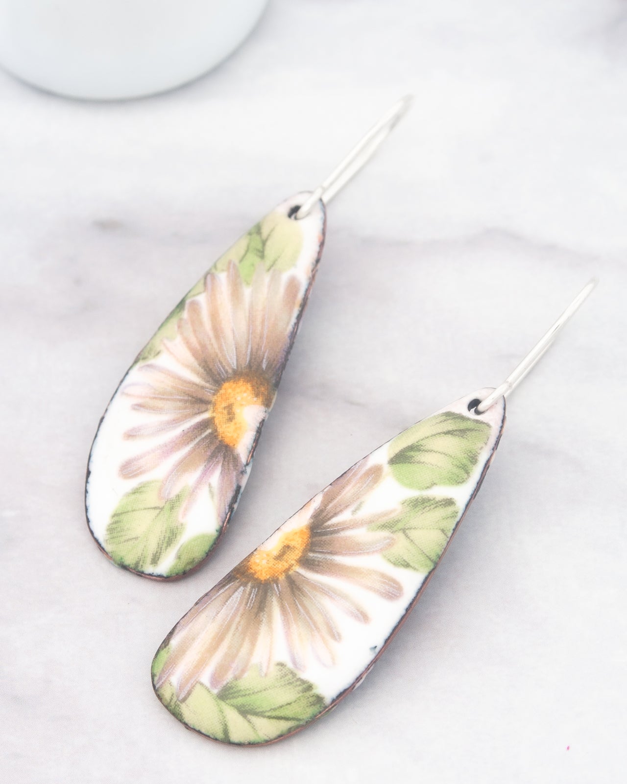 A pair of earrings with a floral design, featuring a large white flower with green leaves and a yellow center.