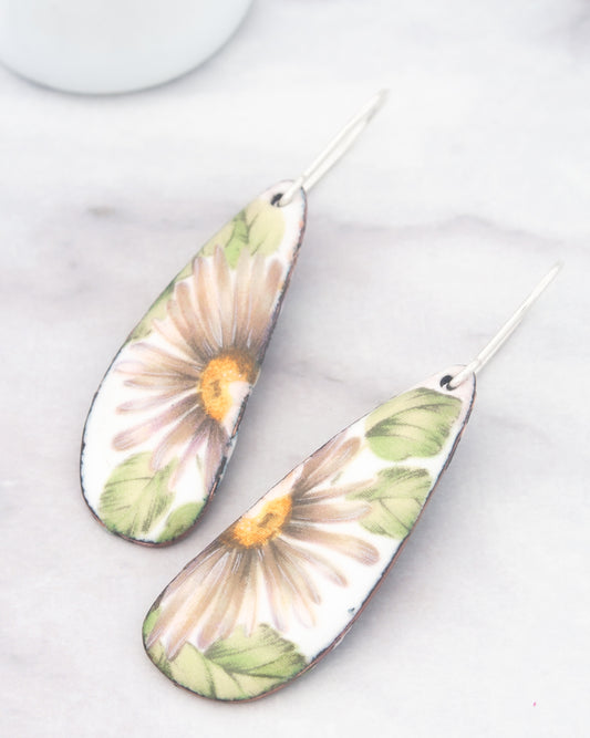A pair of earrings with a floral design, featuring a large white flower with green leaves and a yellow center.