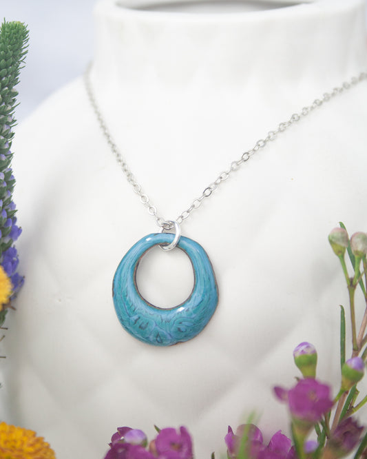 A necklace with a blue enamel pendant is displayed on a white vase, surrounded by colorful flowers.