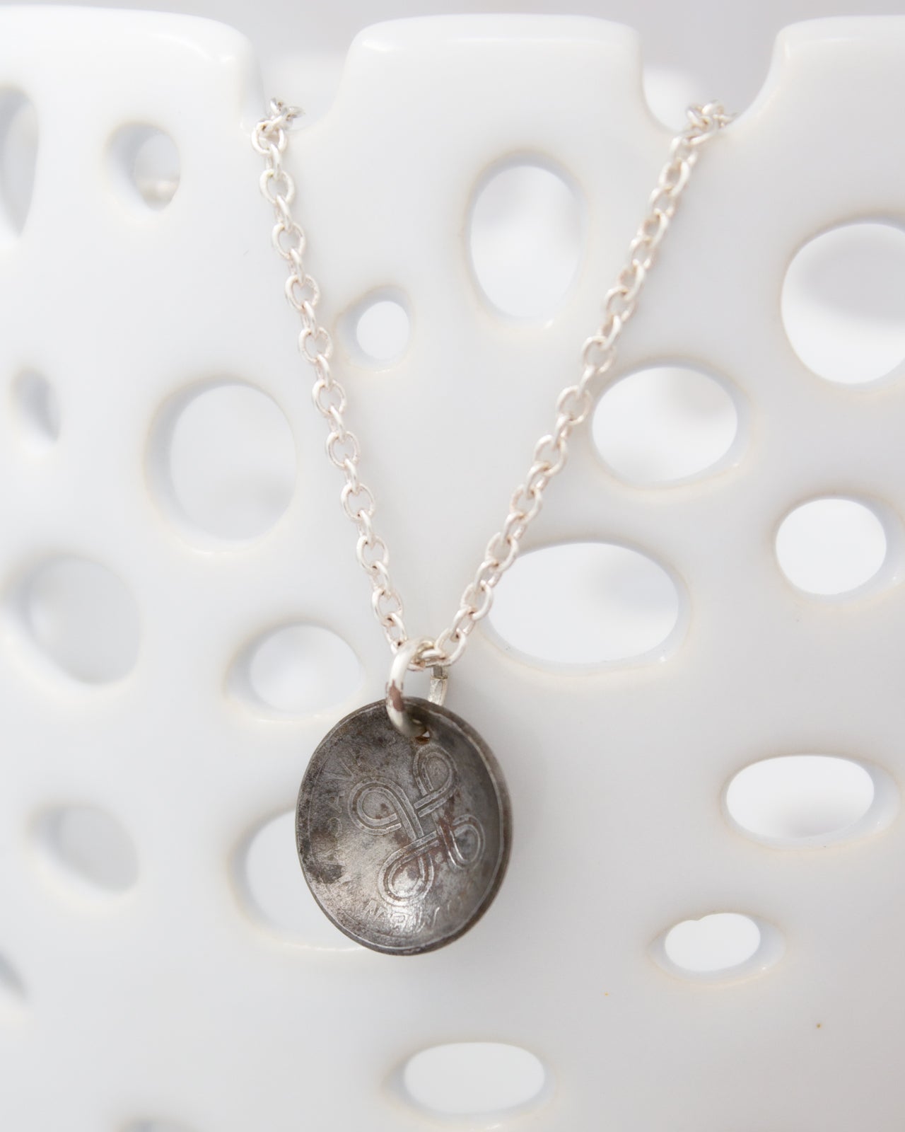 A silver necklace with a pendant featuring a circular design is displayed against a white background with a perforated pattern.