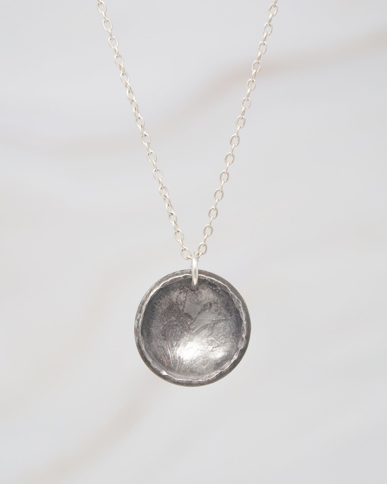 A silver necklace with a round pendant hanging against a white background.