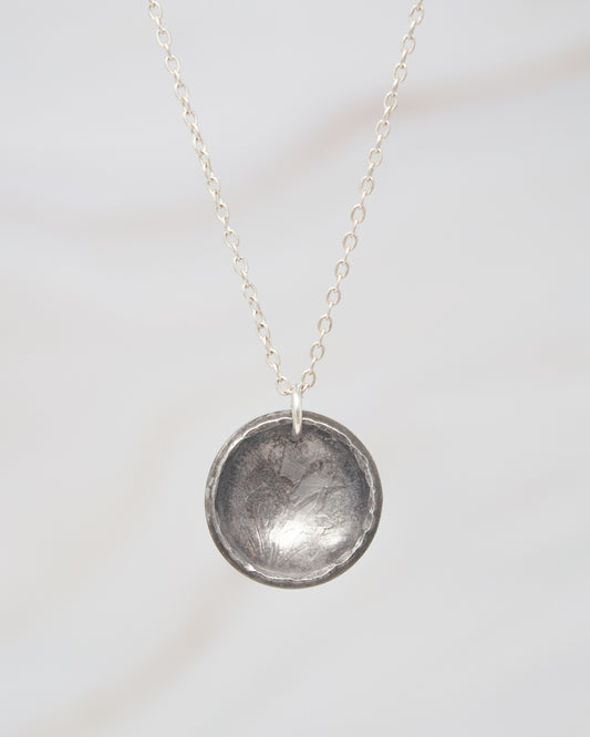 A silver necklace with a round pendant hanging against a white background.
