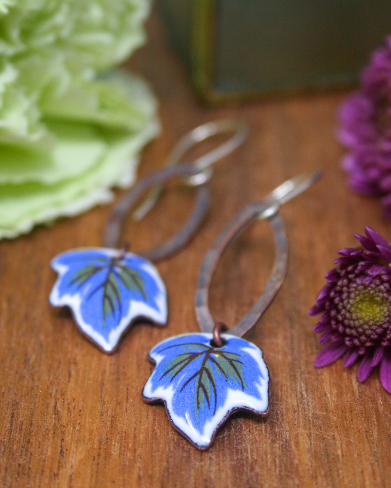 A pair of earrings with blue leaf-shaped designs, placed on a wooden surface alongside some purple flowers.