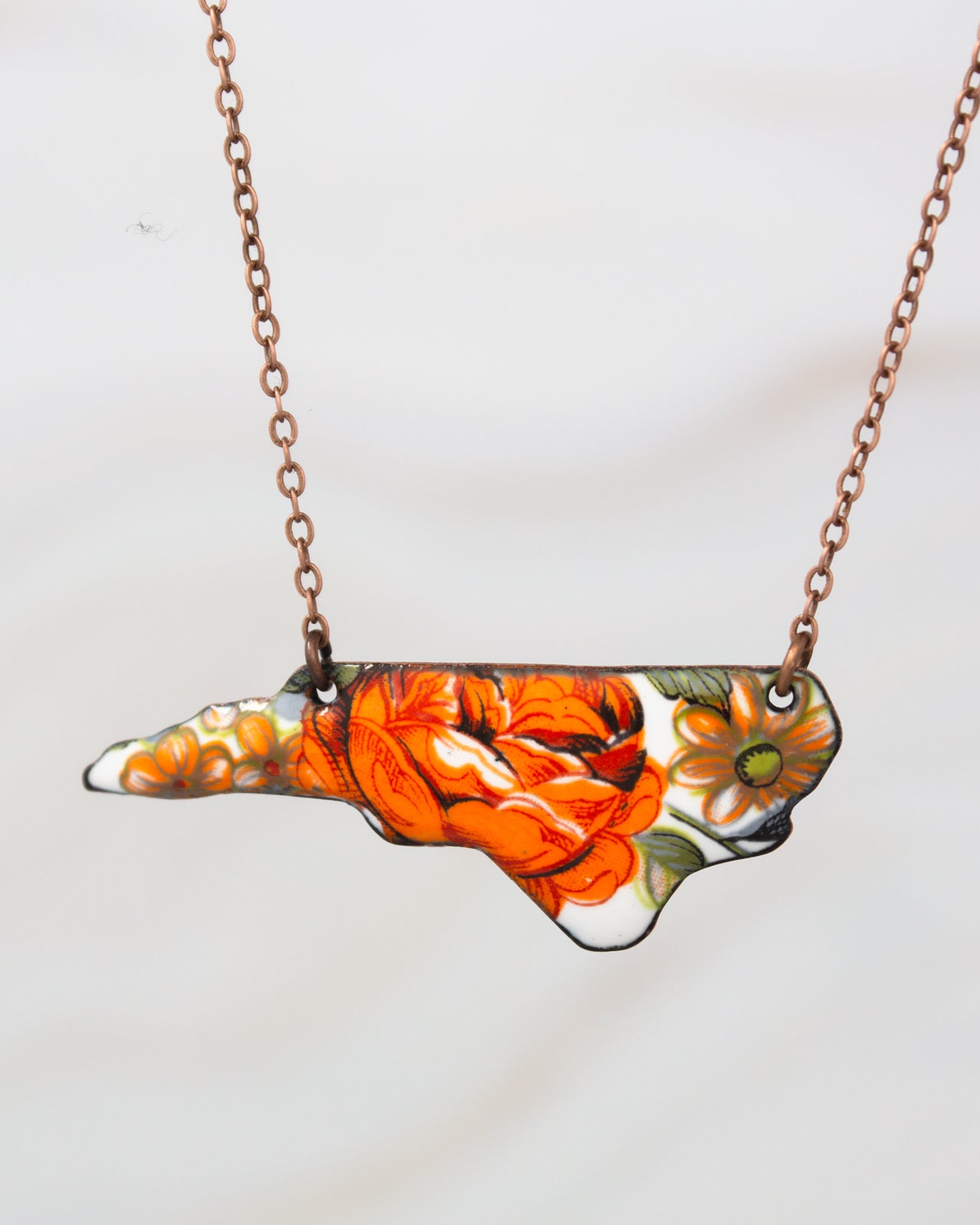 A necklace with a pendant in the shape of the state of North Carolina, featuring a floral design in orange and yellow colors.