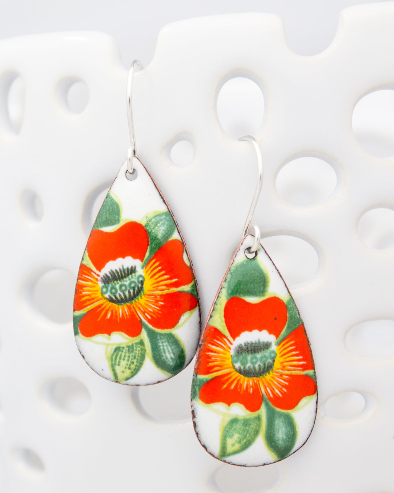 A pair of earrings with a floral design, featuring a large orange flower with green leaves and a smaller red flower with green leaves.