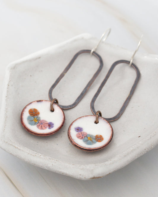 A pair of earrings with floral designs on the circular pendants, displayed on a white plate.