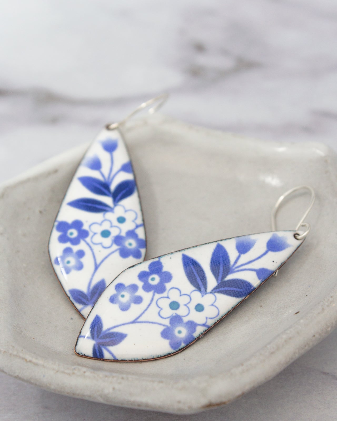A pair of blue and white floral earrings are displayed on a white plate against a marble background.