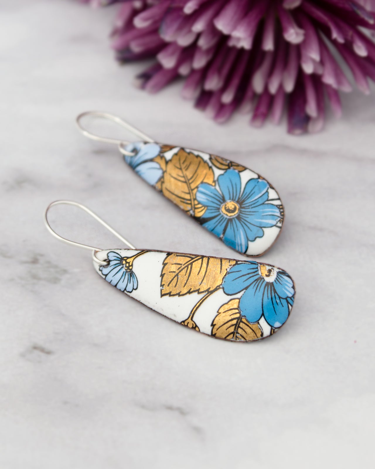 A pair of earrings with a floral design, featuring blue and gold flowers, are displayed on a marble surface alongside a purple flower.