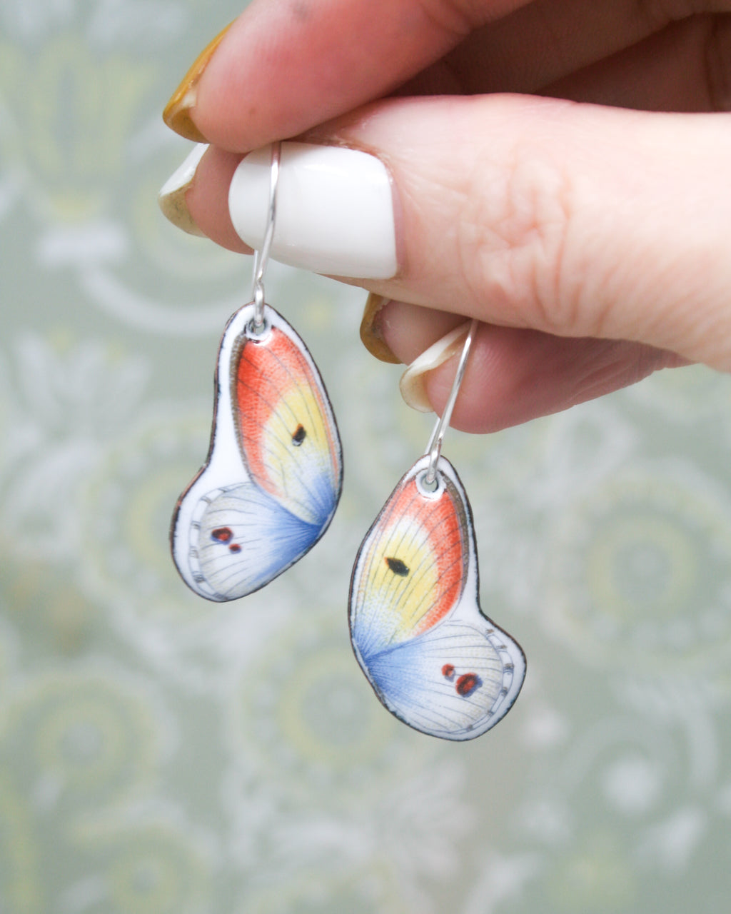 A hand holding a pair of colorful butterfly earrings against a blurred background.