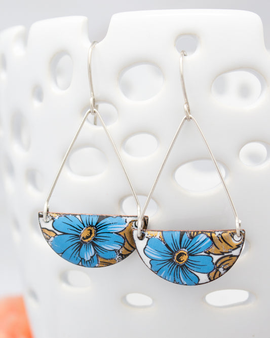 A pair of earrings with blue and gold floral designs hanging from a white ceramic holder.