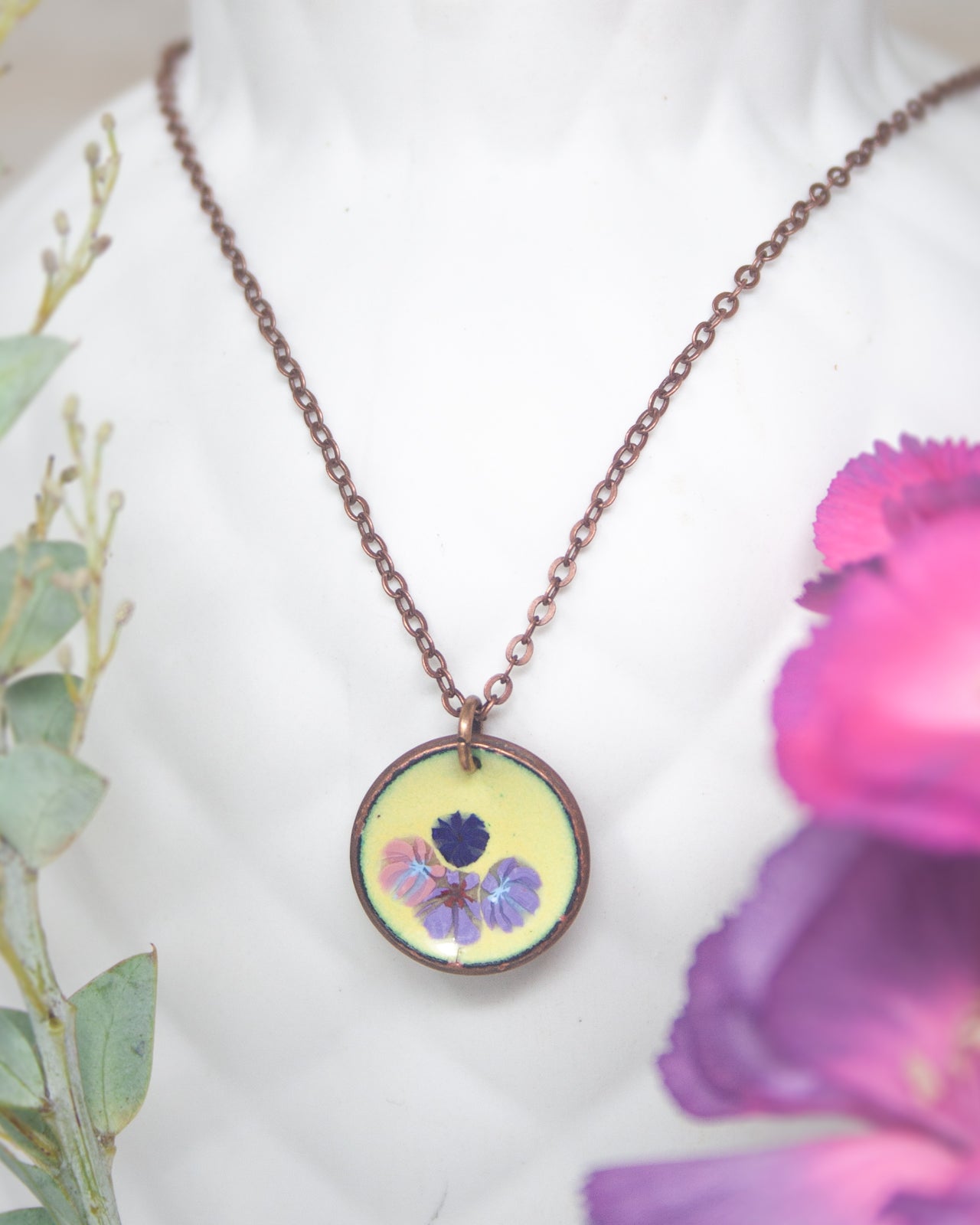 A necklace with a round pendant featuring a floral design, including purple and blue flowers, is displayed against a white background.