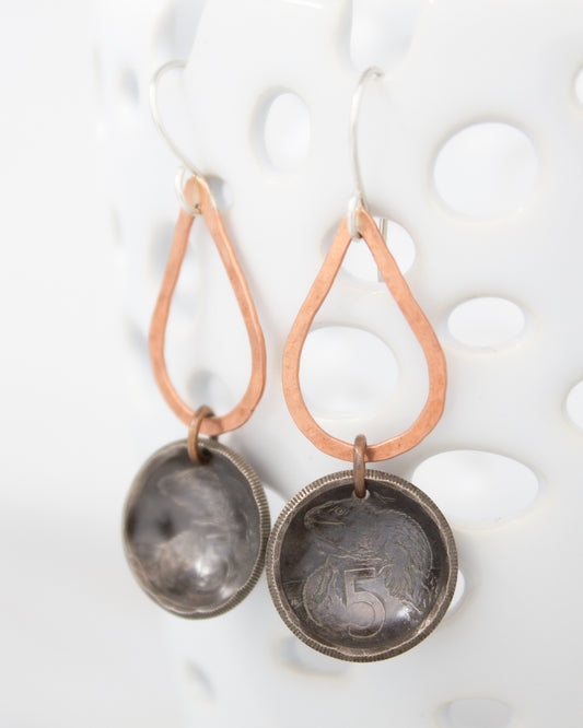 A pair of earrings with a metal pendant and a metal ring hanging from it. The pendant appears to be a small metal coin or token, and the ring is made of metal. The earrings are displayed against a white background, which highlights their design and details.