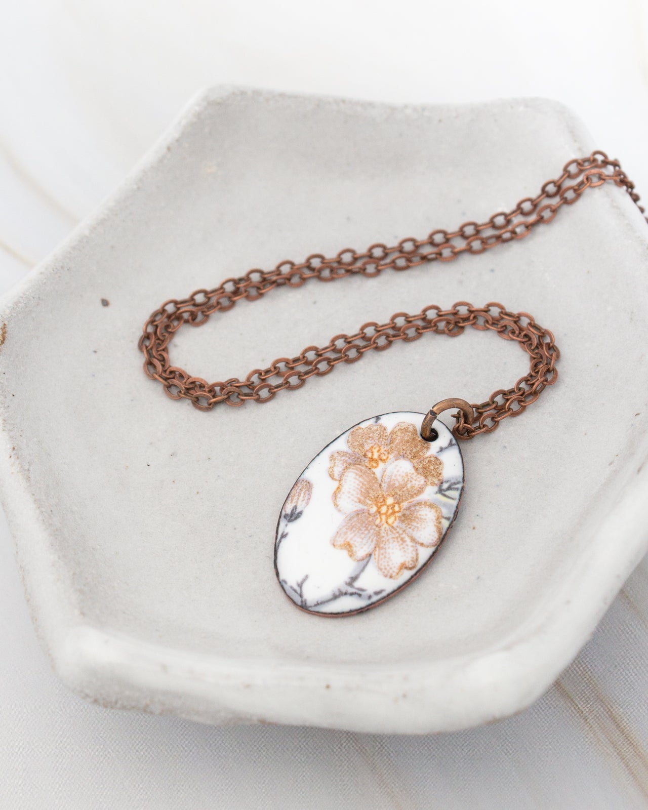 A white ceramic dish containing a brown metal necklace with a pendant featuring a floral design.