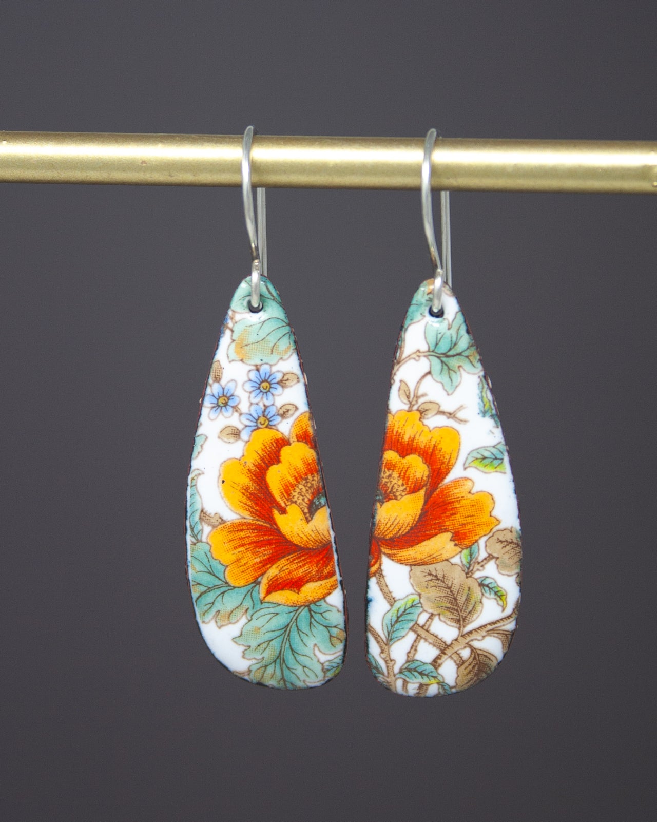 A pair of earrings with a floral design, featuring a combination of orange and white colors, hanging from a gold bar.