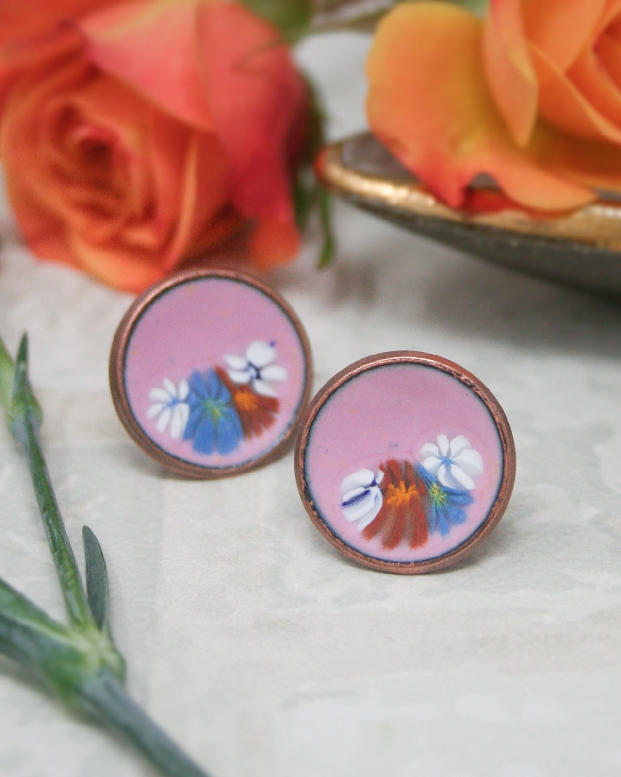 two round earrings with a floral design, placed on a white surface. In the background, there are two roses, one pink and one orange, adding a touch of color to the scene.