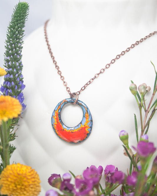 A necklace with a circular pendant featuring a vibrant design of blue and orange colors, surrounded by a variety of colorful flowers and foliage.