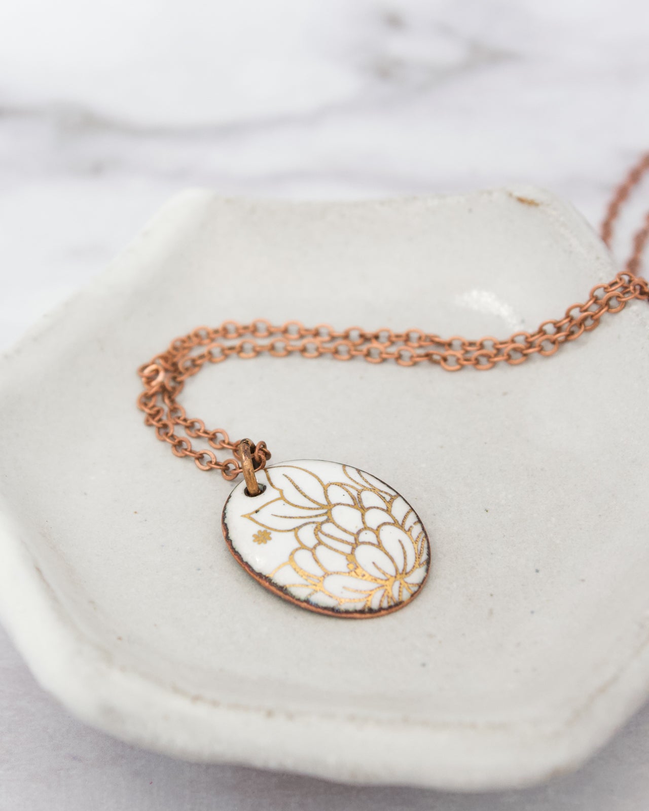 A white ceramic dish containing a delicate necklace with a pendant featuring a floral design in gold and white colors.