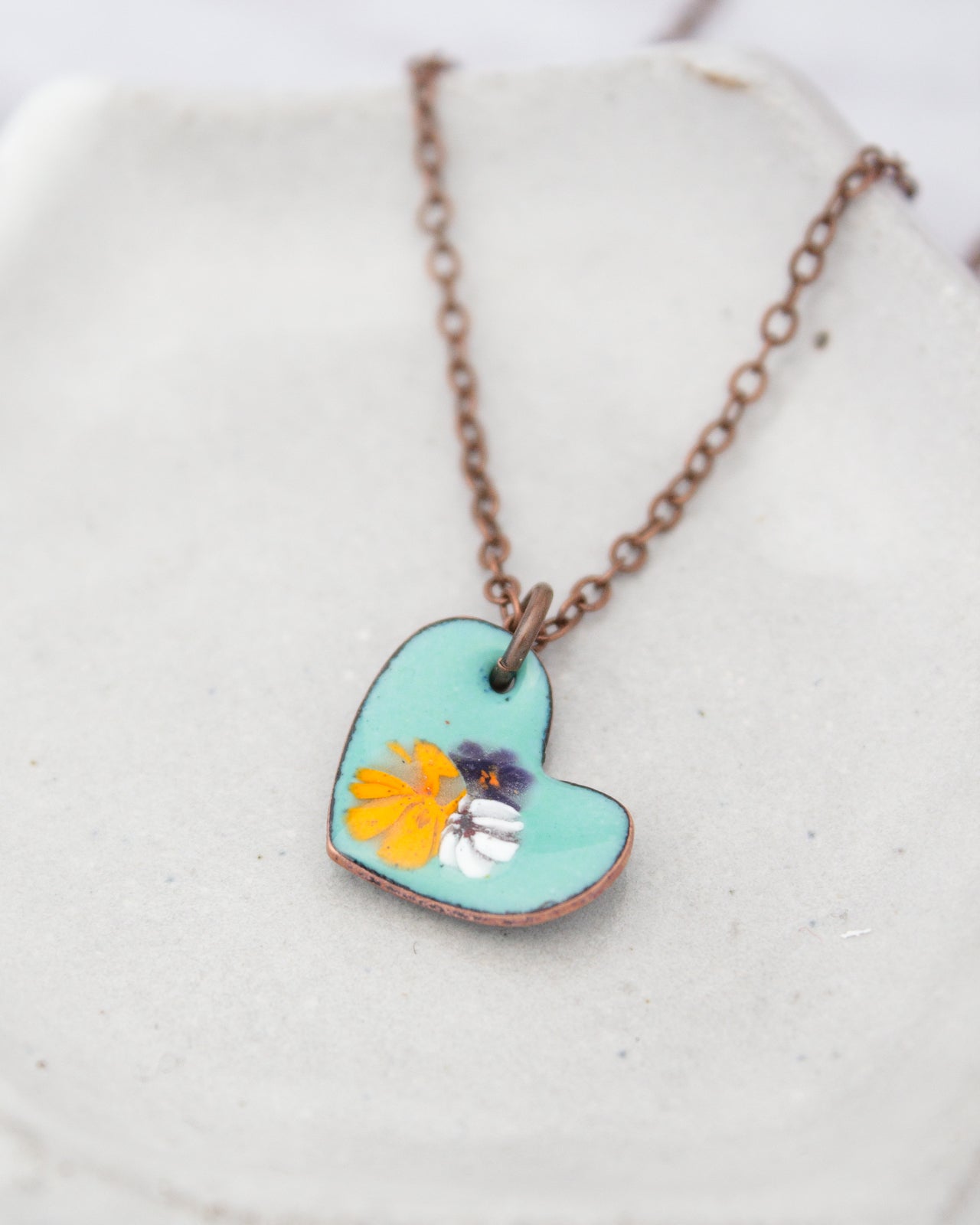 A turquoise heart-shaped pendant with a floral design is displayed on a white surface, attached to a brown chain.
