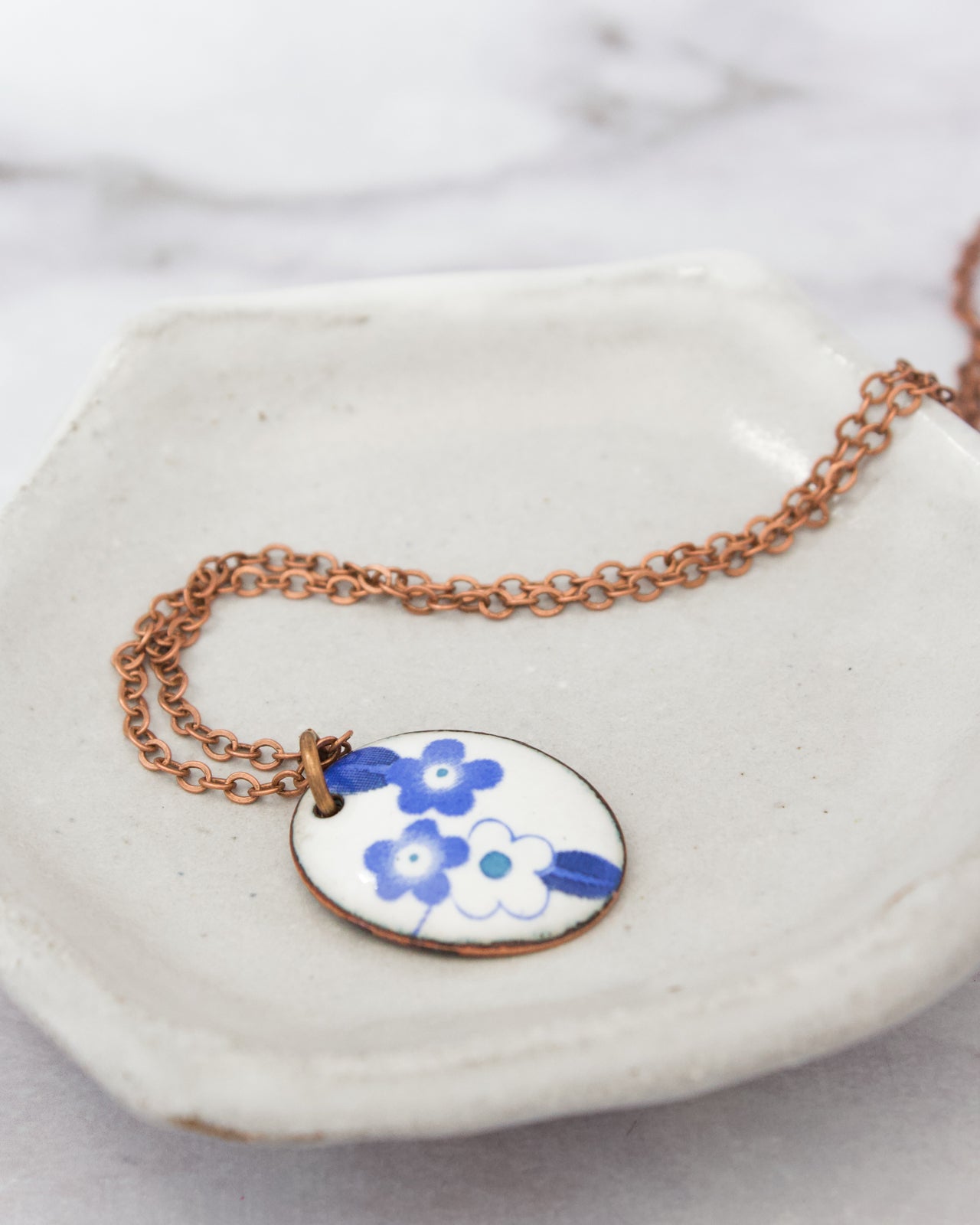A necklace with a pendant featuring blue flowers is displayed on a white plate.