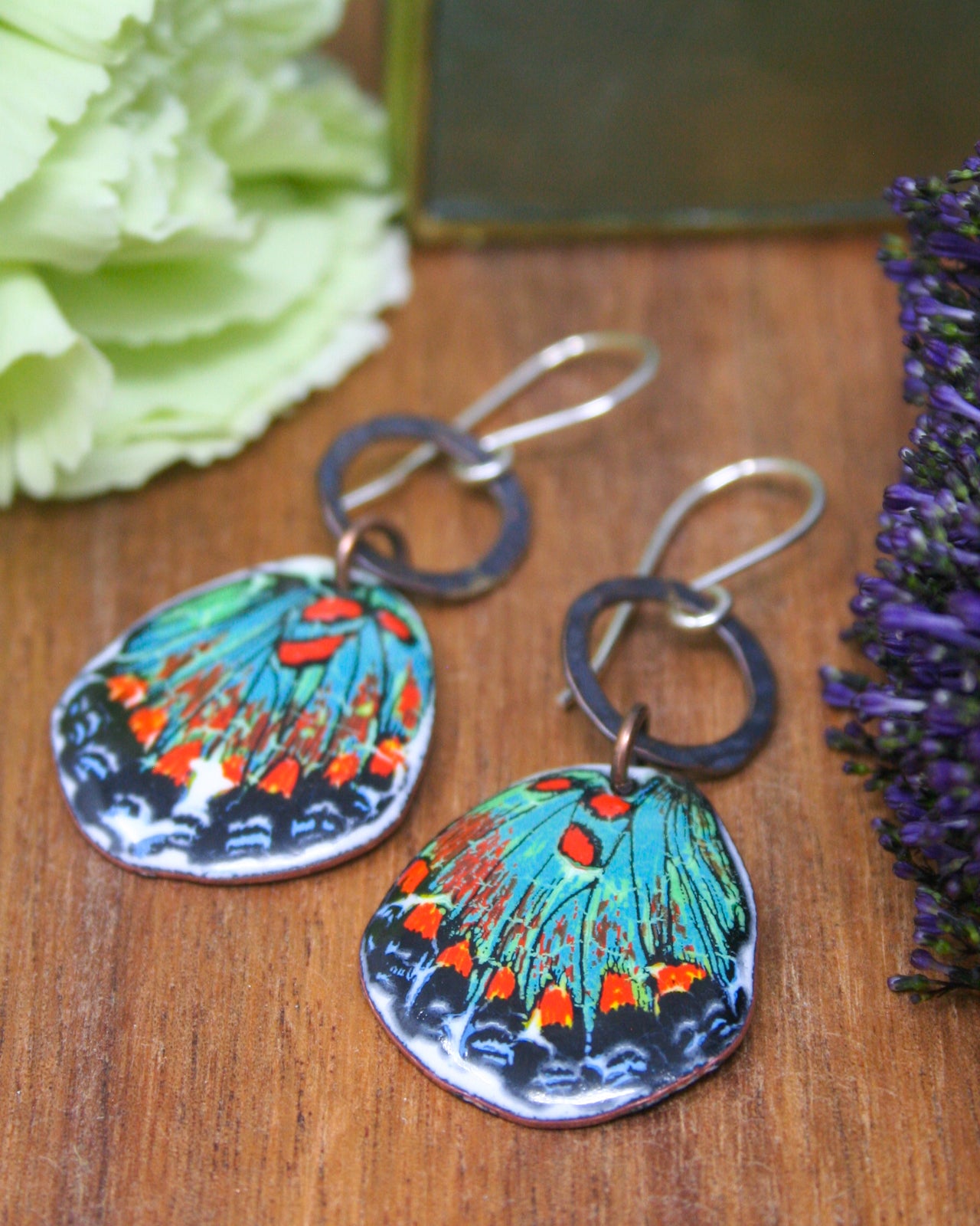 A pair of colorful earrings with butterfly designs, featuring vibrant colors and intricate patterns, are displayed on a wooden surface alongside a green flower.