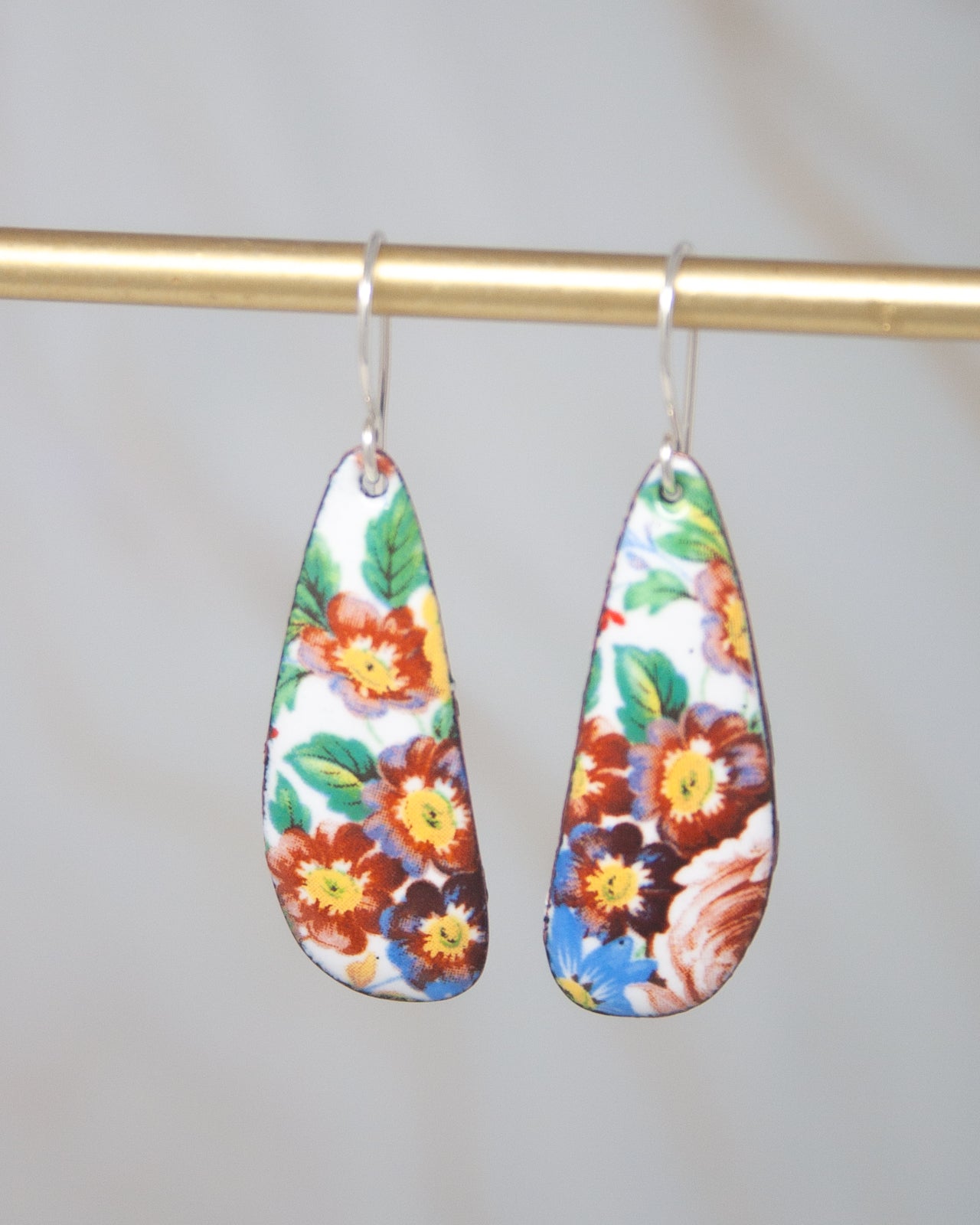 A pair of colorful earrings with floral designs hanging from a gold-colored metal hook.