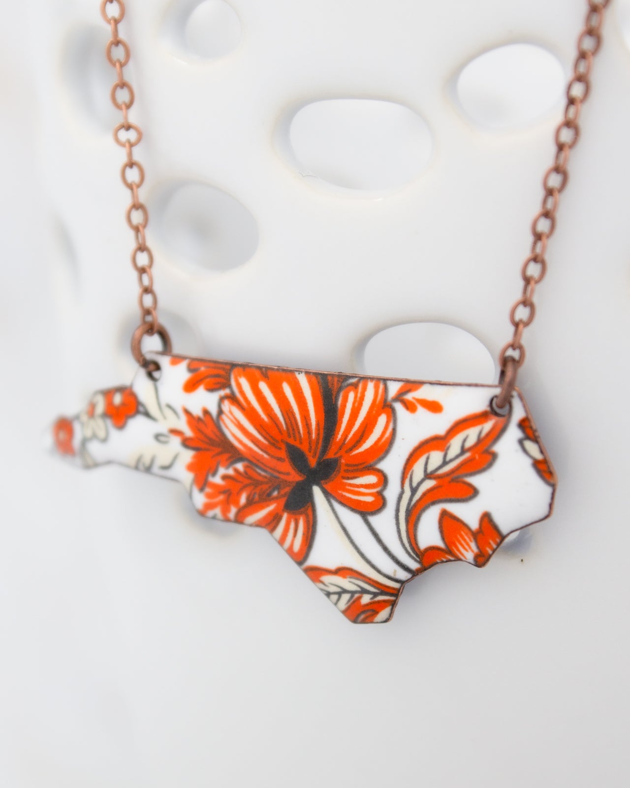 A necklace with a pendant in the shape of the state of North Carolina, featuring a floral design in orange and white colors.