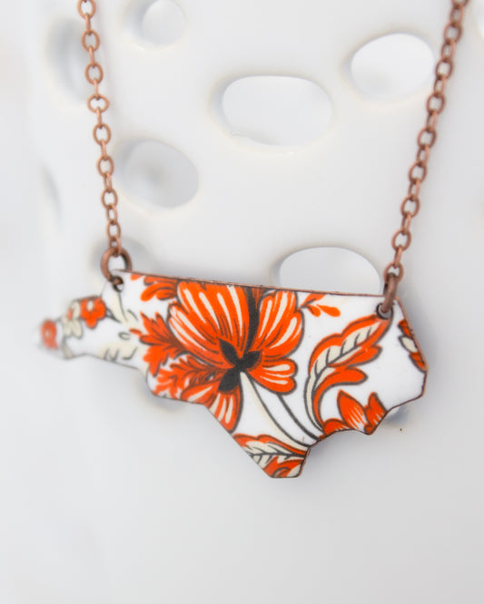 A necklace with a pendant in the shape of the state of North Carolina, featuring a floral design in orange and white colors.