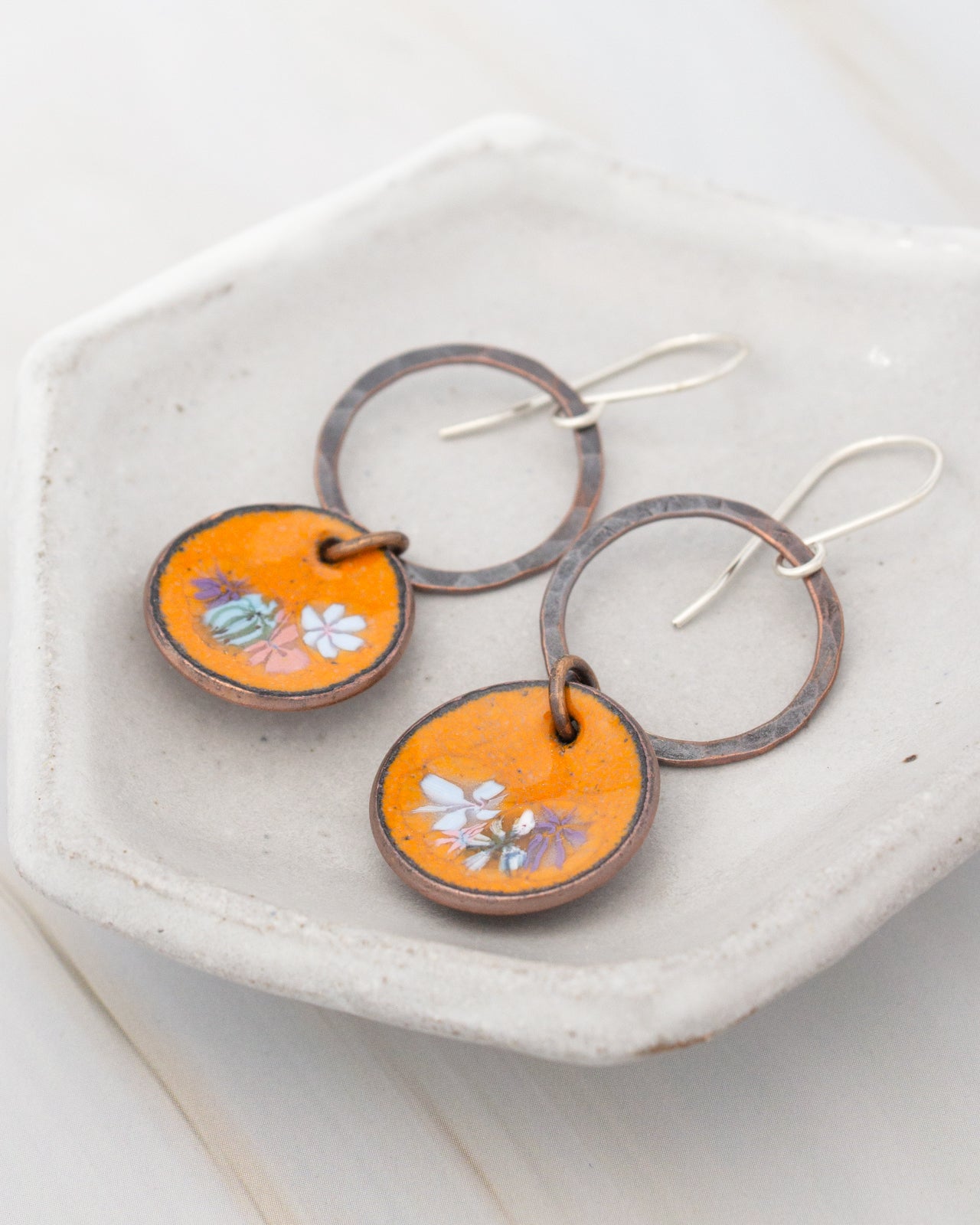 A pair of orange earrings with floral designs on the circular pendants, displayed on a white plate.