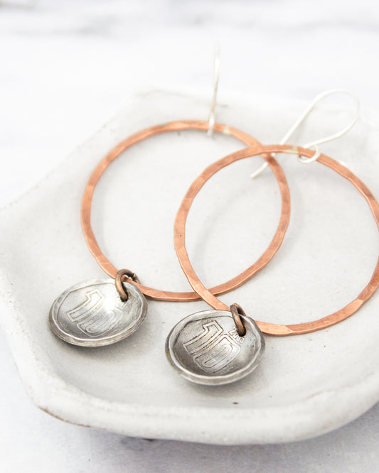 A pair of earrings with a copper-colored metal frame and silver-colored metal pendants, resting on a white surface.