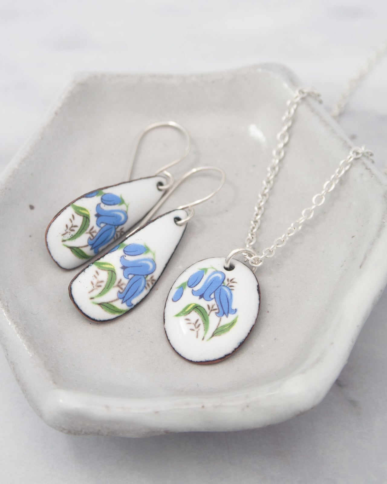 A pair of white earrings with blue floral designs and a necklace with a pendant featuring blue flowers are displayed on a white plate.