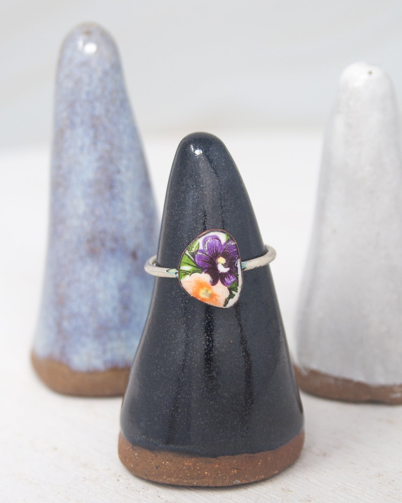 A ring with a purple flower design is displayed on a black stand, with two ceramic or ceramic-like objects in the background.