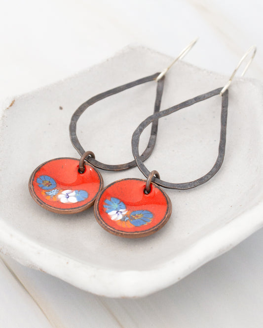 A pair of earrings with red and blue floral designs on circular pendants, resting on a white surface.