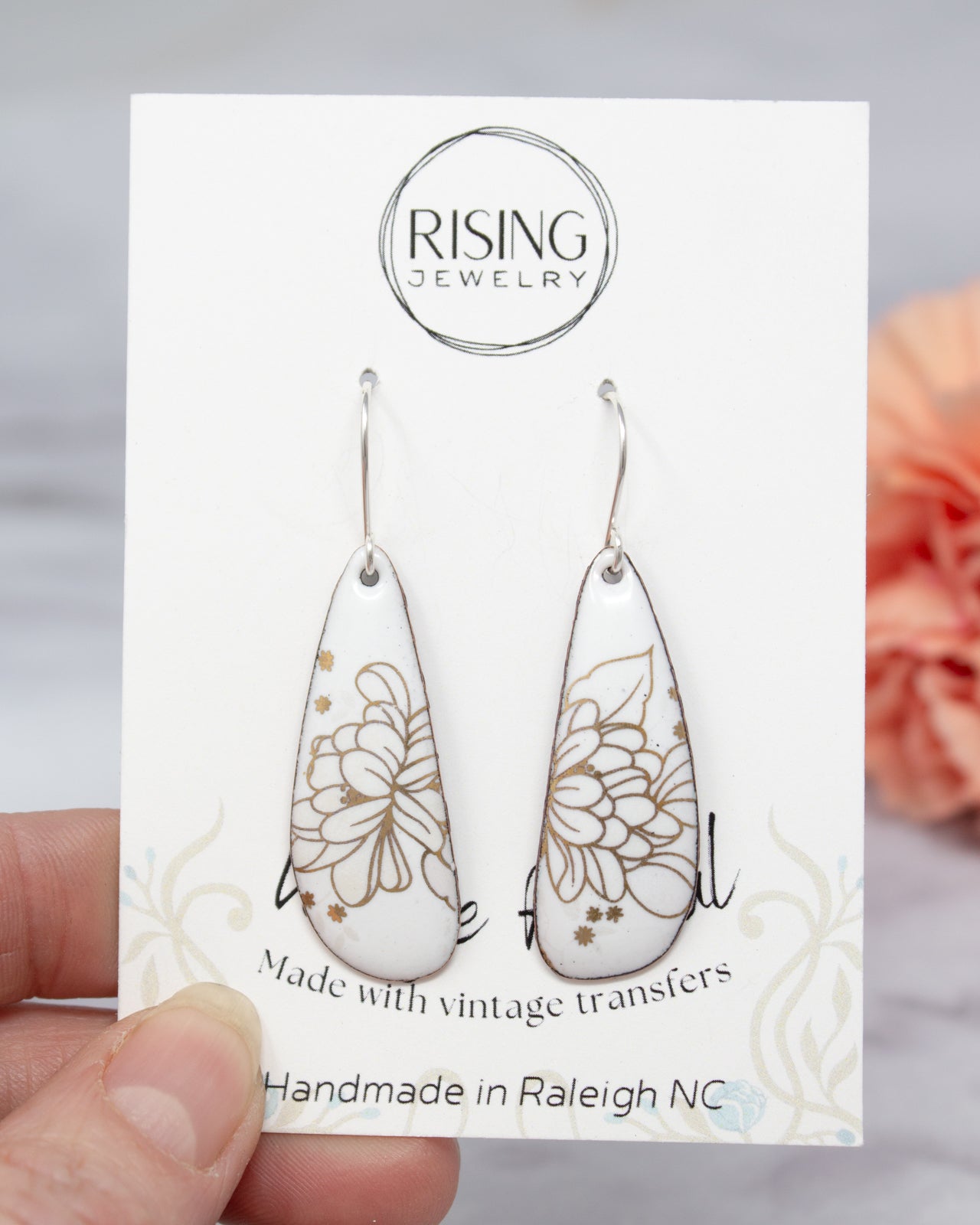 A hand holding a pair of white earrings with gold floral designs, as part of a jewelry product packaging.