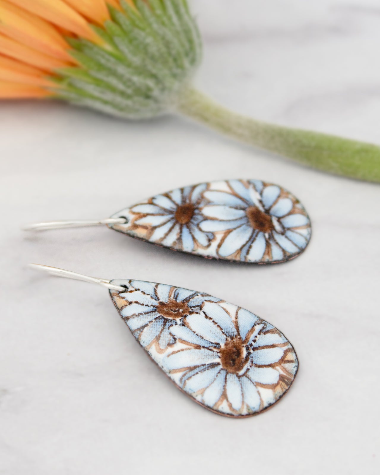 a pair of earrings with blue and white floral designs, placed on a marble surface alongside a green flower.