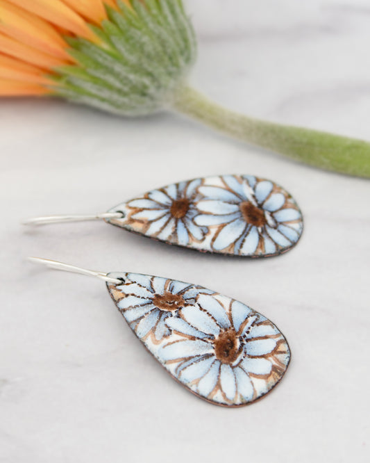 a pair of earrings with blue and white floral designs, placed on a marble surface alongside a green flower.