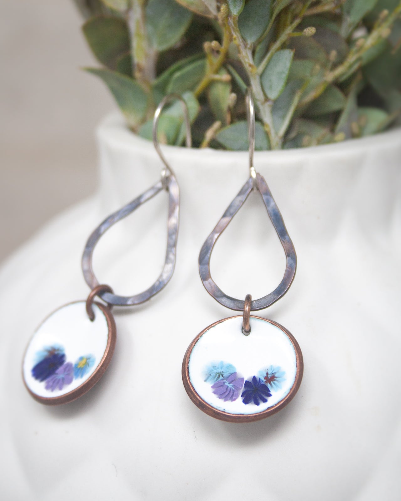 A pair of earrings with floral designs on the circular pendants, hanging from a white vase with greenery.