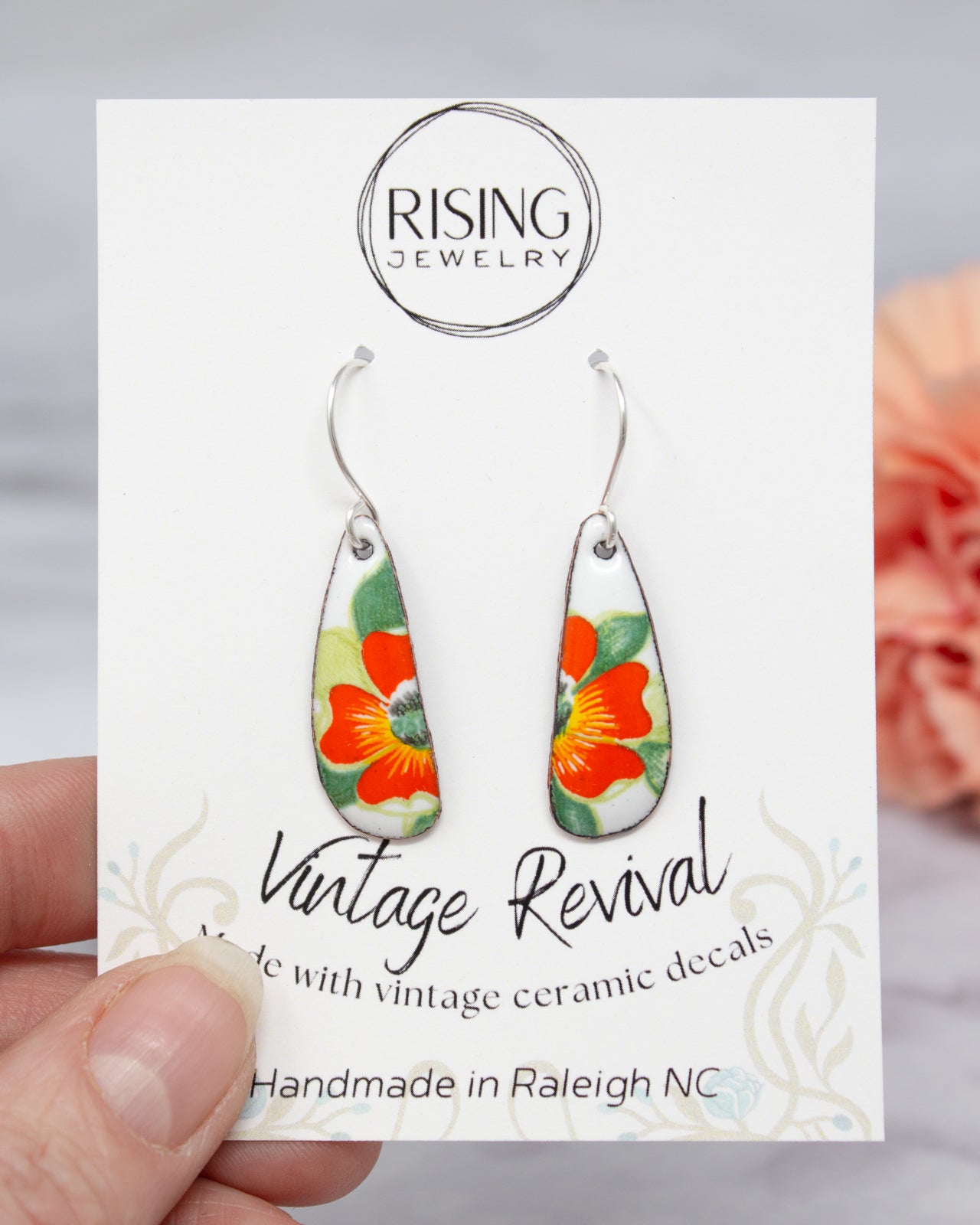 A hand holding a pair of earrings with a floral design, against a blurred background.