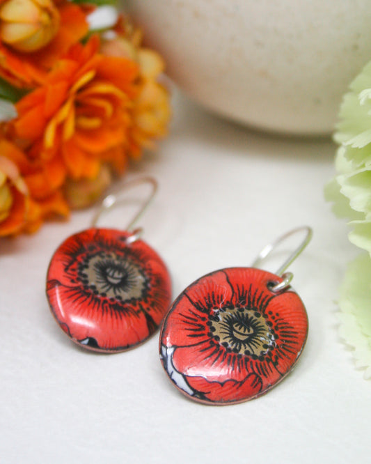 a pair of red earrings with a floral design, placed on a white surface next to a bouquet of orange and yellow flowers.