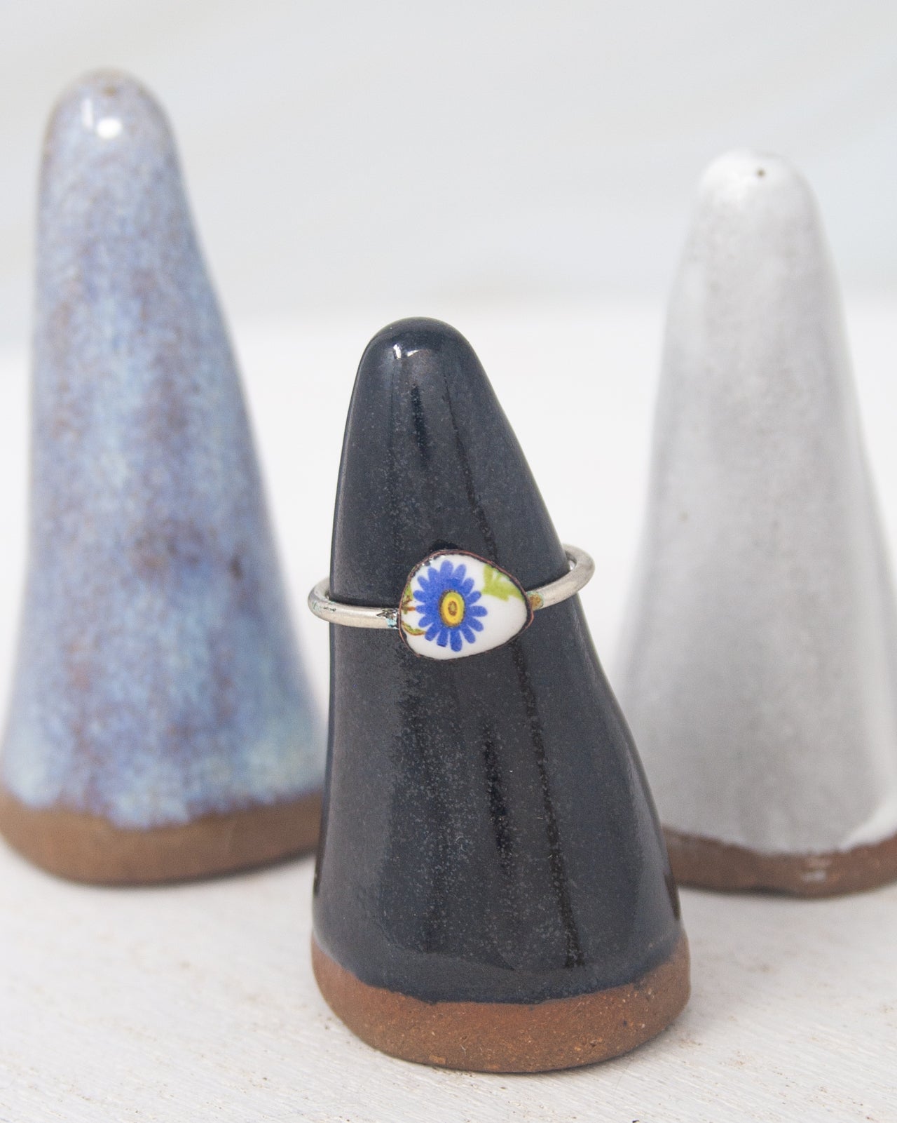 A ring with a blue flower design is displayed on a black stand, with two other ceramic stands in the background.