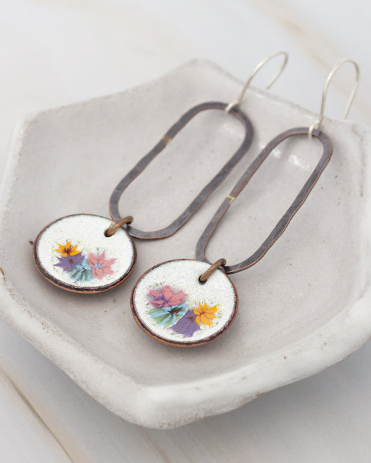 A pair of earrings with floral designs on the circular pendants, displayed on a white plate or tray.