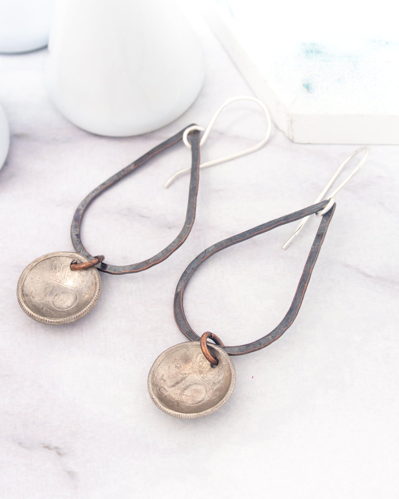 A pair of earrings with a metal frame and a metal coin charm hanging from them, placed on a white marble surface.